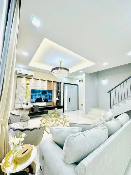 Living features: light grey fabric sofa, gold coffee table, chandelier, recessed ceiling lights, wall-mounted TV, stone feature wall, white painted walls, cream tiled flooring, curtains, air conditioning unit, staircase with metal railing; style: modern.