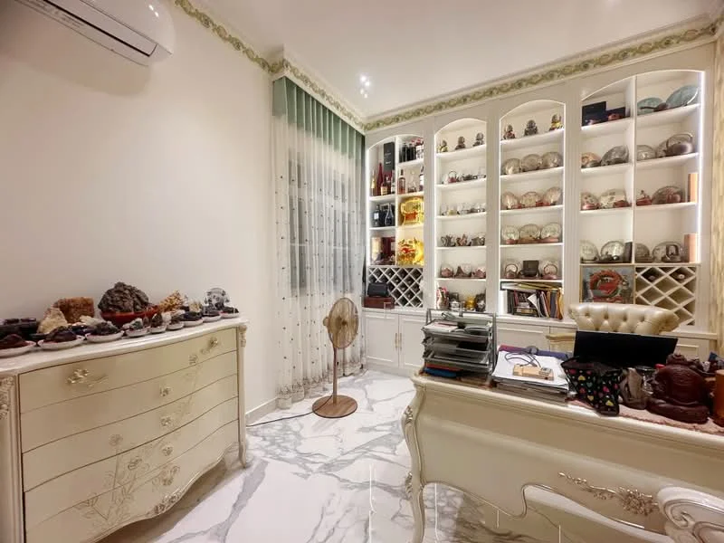 Study features: white painted walls, marble flooring, built-in shelving unit with glass doors, decorative items on shelves, desk with ornate design, white upholstered chair, ceiling-mounted air conditioning unit, floor fan, curtains with green accents, decorative crown moulding; style: traditional.