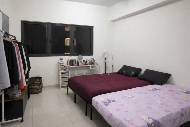 Bedroom features: two single beds with purple floral bedding, white painted walls, glossy tile flooring, black framed windows, clothes rack with hanging clothes, white shelving unit with various items, small fan, decorative basket, nightstand with personal items.