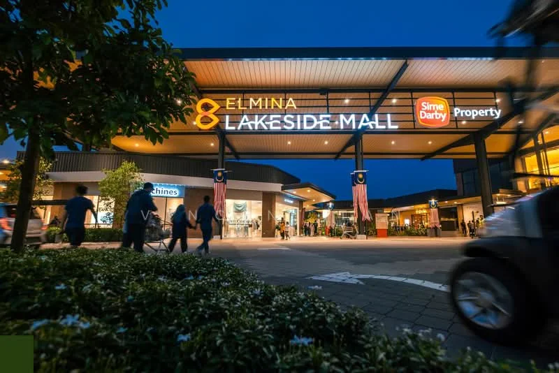 Exterior type: shopping mall; features: modern facade with glass and metal, illuminated signage, wide entrance, landscaped garden with shrubs, paved walkway, outdoor seating area, decorative flags, multiple storefronts, nighttime lighting, open-air design; style: modern.