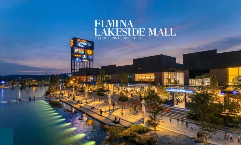 Exterior type: shopping mall; features: modern facade with glass and metal cladding, multiple storeys, large illuminated signage, surrounded by water, landscaped areas with trees, outdoor walkway, people walking along the waterfront, evening lighting, reflective water surface, parking area visible; style: contemporary.