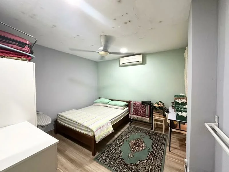 Bedroom features: queen-size bed with green bedding, wooden bed frame, ceiling fan, wall-mounted air conditioning unit, light green painted walls, laminate flooring, decorative area rug with intricate pattern, built-in wardrobe, small bedside table, stacked blankets on shelf.