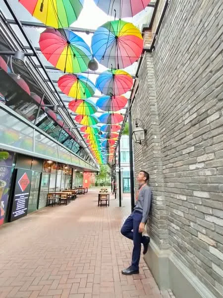 Exterior features: brick facade, colorful umbrellas hanging overhead, paved walkway, outdoor seating area with tables and chairs, glass storefronts, decorative wall lighting; style: modern.