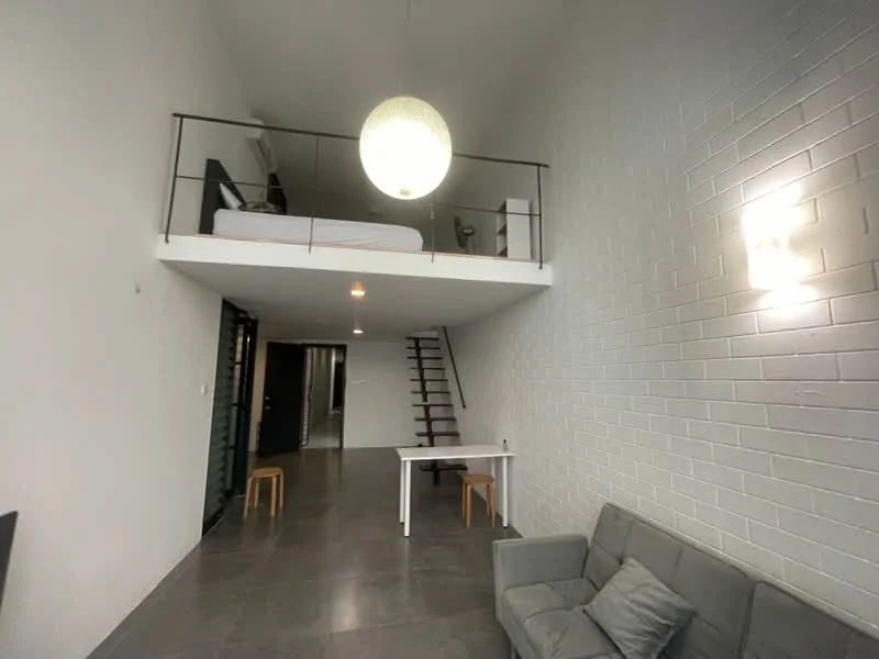 Living features: grey fabric sofa, white painted walls, exposed brick wall, concrete flooring, pendant light fixture, staircase with wooden steps, small dining table, two wooden stools; style: modern. Room features: lofted sleeping area with bed, wall-mounted air conditioning unit.