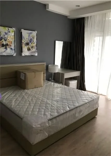 Bedroom features: queen-size bed with mattress protector, grey accent wall, wooden flooring, two framed artworks on wall, dressing table with mirror, table lamp, curtains, natural light from large window; style: modern.