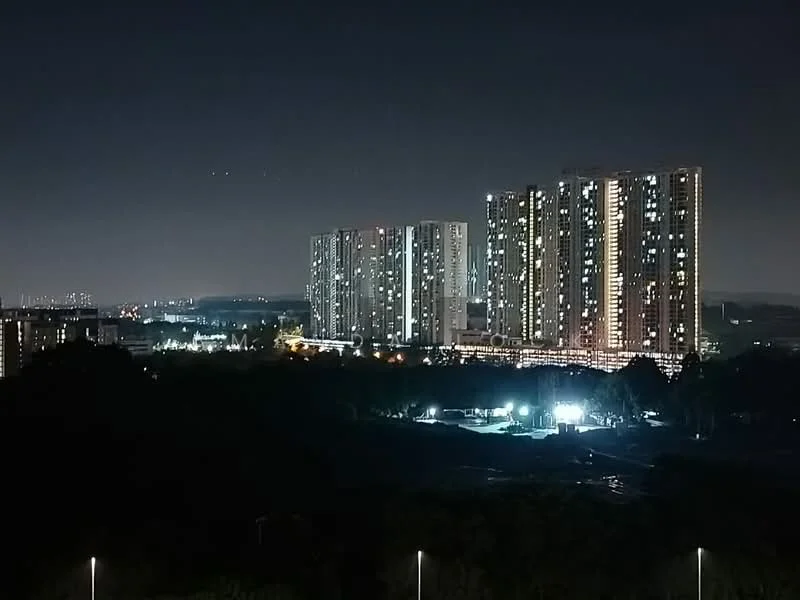 Exterior type: high-rise condominium; features: multiple high-rise buildings, illuminated facade, glass curtain wall, surrounded by greenery, nighttime city view, parking area with lights, landscaped grounds; style: modern.