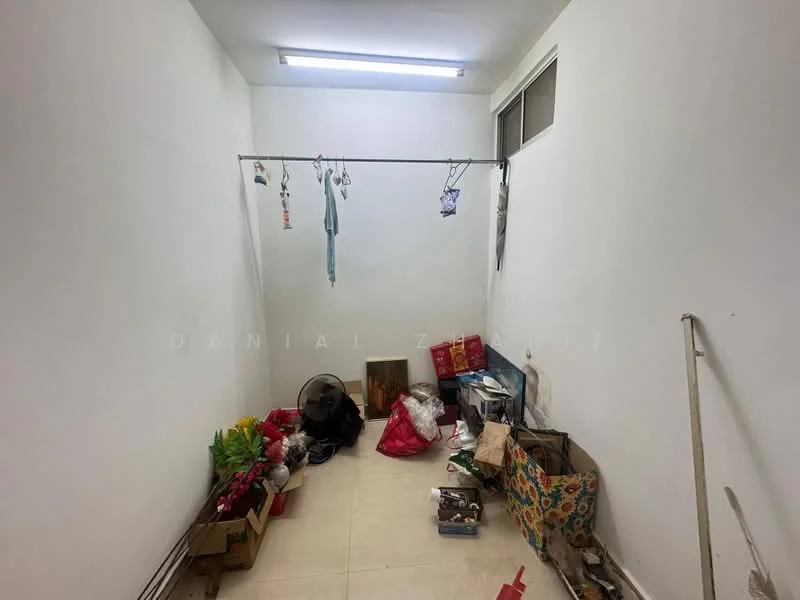 Room features: white painted walls, tile flooring, overhead fluorescent lighting, clothes hanging on a rod, fan, various boxes and bags, houseplants in pots, cluttered space.