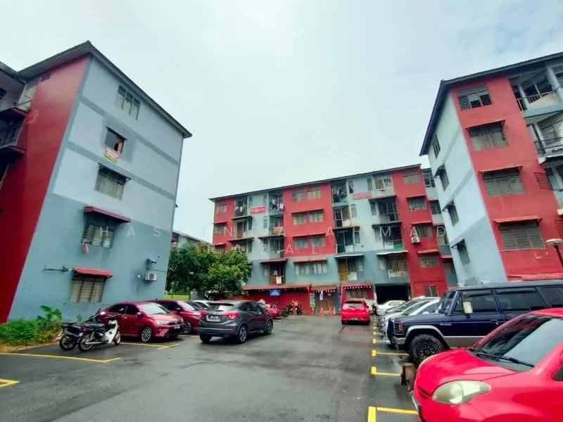 Exterior type: low-rise apartment block; features: red and blue painted facade, 2-storey building, parking lot with marked spaces, surrounded by greenery, balconies on upper floors, multiple vehicle parking, visible air conditioning units, windows with metal grilles; style: modern.