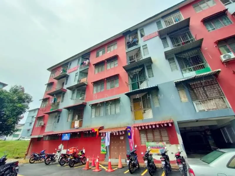 Exterior type: low-rise apartment block; features: red and blue painted facade, multiple storeys, balconies with metal railings, window grilles, visible air conditioning units, parking area with motorcycles, decorative banners, landscaped grassy area, entrance with cones, surrounding greenery; style: modern.