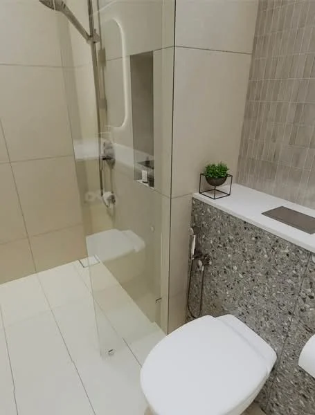 Bathroom features: walk-in shower with glass enclosure, chrome handheld shower head, white toilet, grey stone wall cladding, white countertop, small potted plant, cream tiled walls, light-colored floor tiles; style: modern.