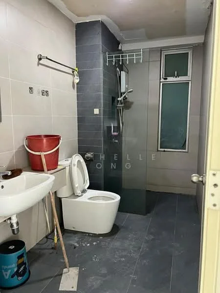 Bathroom features: white ceramic toilet, single-sink vanity with wall-mounted faucet, shower area with glass enclosure, grey wall tiles, light grey floor tiles, chrome shower head, towel rack, window for natural light, bucket, cleaning supplies, plumbing fixtures visible.