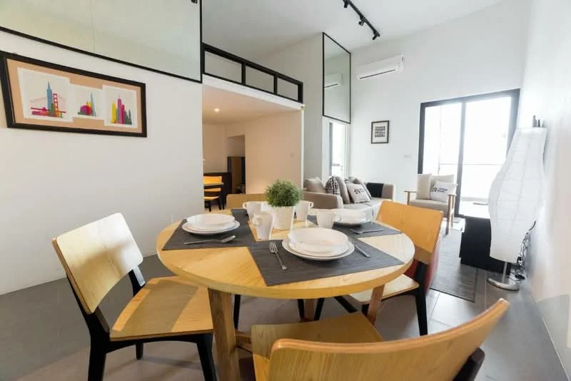Dining features: round wooden dining table, 4 wooden dining chairs, table setting with plates and cups, grey table runner, decorative plant in a pot, wall-mounted artwork with cityscape, light-colored walls, floor-to-ceiling windows, air conditioning unit on wall, laminate flooring; style: modern. Living features: sofa, coffee table, area rug, floor lamp, wall-mounted artwork, light-colored walls, floor-to-ceiling windows; style: modern.