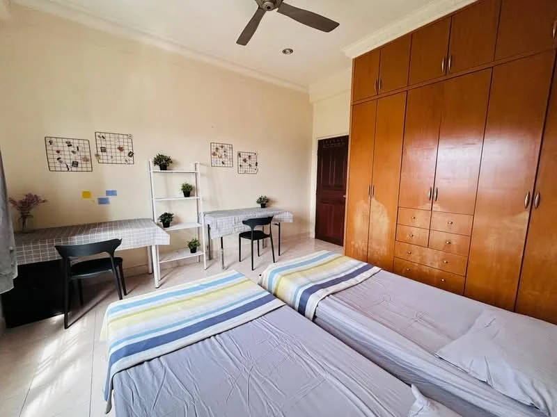Bedroom features: two single beds with light grey bedding, wooden wardrobe with multiple drawers, study desk with white table and black chair, light-colored walls, ceiling fan, decorative wall art, small potted plants on shelves, window with sheer curtains; style: modern.