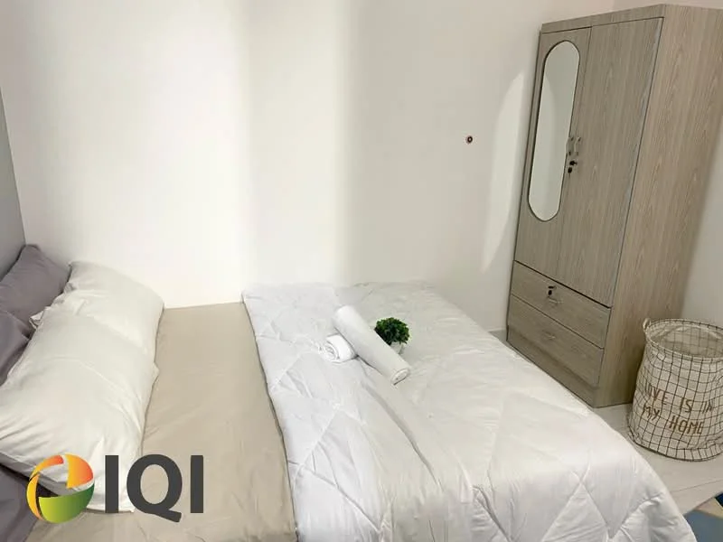 Bedroom features: queen-size bed with beige bedding, white quilted comforter, grey pillow, wooden wardrobe with mirror, white decorative towel, small potted plant, woven laundry basket, plain white walls, laminate flooring; style: minimalist.