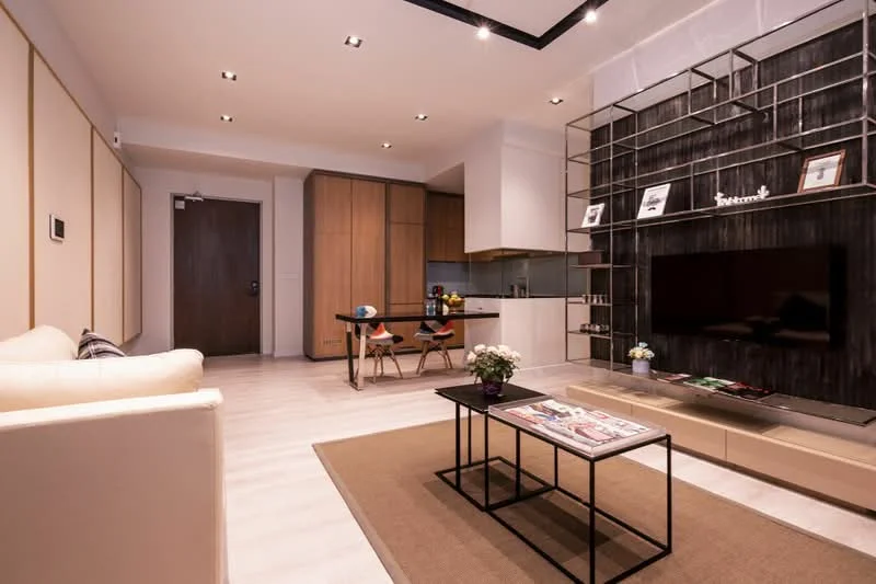 Living features: white painted walls, light wood flooring, recessed downlights, ceiling with integrated lighting, built-in shelving unit with dark finish, wall-mounted TV, coffee table with black frame, area rug with neutral color, sofa in cream fabric, decorative plants, magazines on coffee table; style: modern. Kitchen features: light wood cabinetry, white countertop, stainless-steel appliances, open-plan layout, bar stools at kitchen island; style: modern.