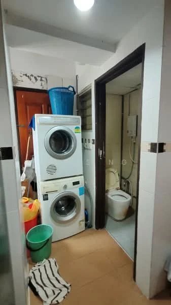 Laundry features: front-load washing machine, tumble dryer, toilet, white tiled walls, grey tiled flooring, basket for laundry, plastic bucket, striped floor mat, wall-mounted water heater, storage shelves, light fixture on ceiling.