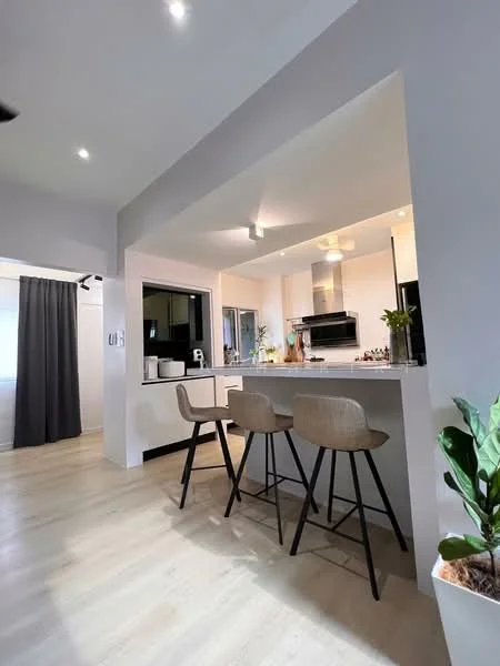 Kitchen features: white painted walls, light wood flooring, recessed downlights, kitchen island with seating, stainless-steel microwave, built-in oven, stainless-steel range hood, black framed window, potted plant, grey curtains, countertop with bar stools; style: modern.