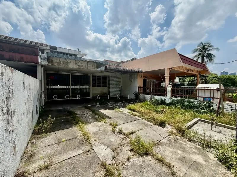 Exterior type: detached bungalow; features: concrete patio, overgrown grass, partially covered outdoor area, single-storey structure, surrounding greenery, adjacent building, cloudy sky, urban context; style: traditional.