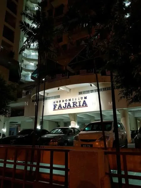 Exterior type: mid-rise condominium; features: illuminated building sign, multiple floors visible, parking area with vehicles, surrounding greenery, balconies on upper floors, entrance with awning, decorative fencing in foreground; style: modern.