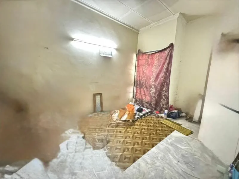Room features: plain white walls, textured ceiling, window with sheer curtain, brown patterned flooring, decorative rug, folded fabric draped over window, scattered personal items, light fixture on wall.
