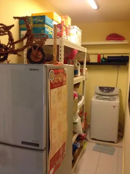 Laundry features: white top-load washing machine, silver refrigerator, shelving unit with storage boxes, tile flooring, yellow painted walls, overhead lighting, decorative wooden bicycle, cleaning supplies on shelves, laundry area with space for storage.