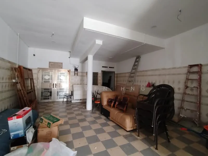 Room features: checkered tile flooring, white painted walls, exposed ceiling, two metal doors, wooden ladder, plastic chairs, brown leather sofa, cardboard boxes, various items scattered, wall-mounted light fixtures.