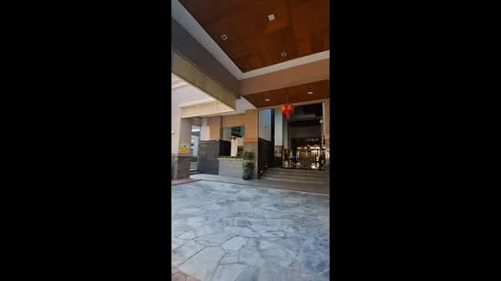 Exterior type: main entrance gate; features: stone-paved walkway, glass entrance doors, decorative lantern, wooden ceiling, planters with greenery, steps leading to entrance, modern facade, overhang roof; style: modern.