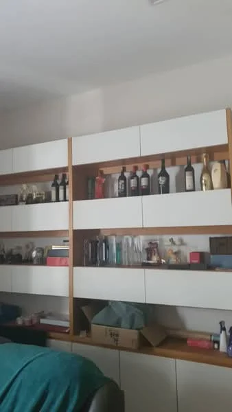 Room features: built-in shelving unit, white cabinet doors, wooden shelving, various bottles displayed, decorative items on shelves, green fabric throw, boxes on lower shelf, neutral wall color; style: modern.