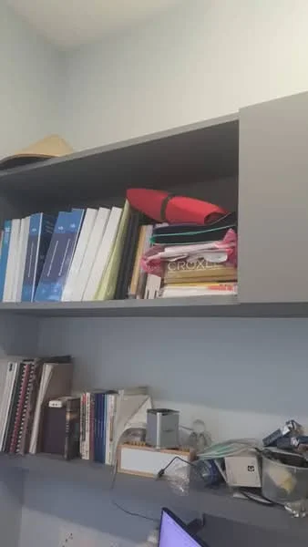 Study features: grey shelving unit, stacked binders, red folder, various books, paperwork, desk with clutter, light blue painted walls.