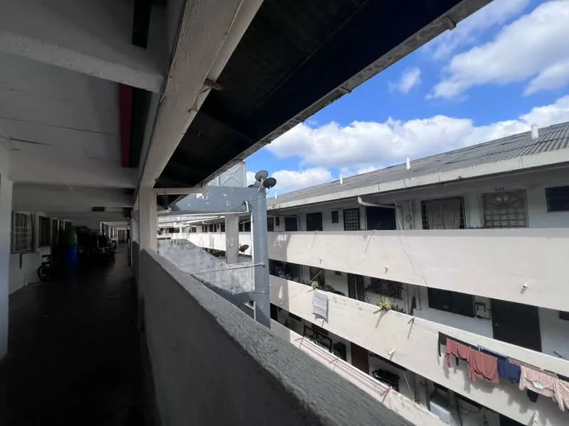 Exterior type: low-rise apartment block; features: concrete facade, two-storey building, balconies visible, laundry hanging on balconies, metal roof, open corridor, surrounding greenery, clear blue sky; style: modern.