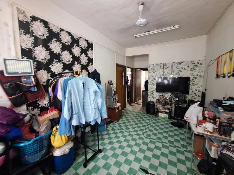Room features: green and white checkered flooring, floral wallpaper, plain white walls, ceiling fan, wall-mounted TV, wooden door, clothing rack with hanging clothes, plastic storage baskets, decorative medals on display, various items on the floor.