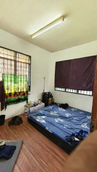 Bedroom features: wooden flooring, white painted walls, single bed mattress on blue bed frame, window with metal grille, blackout curtains, ceiling light fixture, various personal items on floor, small bedside table, clothes on the floor.