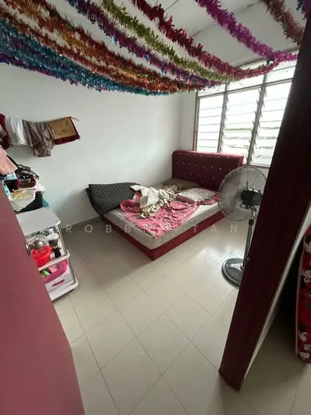 Bedroom features: single bed with mattress and bedding, decorative wall with tinsel garland, white painted walls, floor tiles, window with metal grilles, standing fan, small bedside table, clothing hanging on wall.