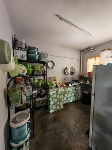 Kitchen features: green patterned countertop, stainless-steel gas stove, refrigerator, metal shelving unit, various green bowls and containers, cooking utensils, sink, exposed pipes, concrete flooring, wall-mounted light fixture, window with sheer curtain.
