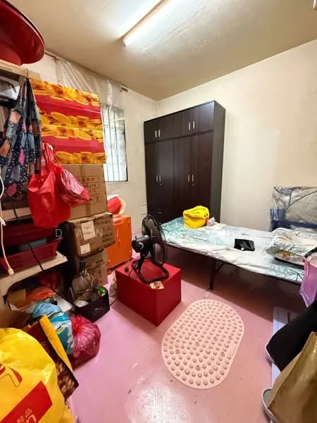 Room features: pink vinyl flooring, white walls, wooden wardrobe, single bed with mattress, fan, storage boxes, shelves with bags, decorative mat.