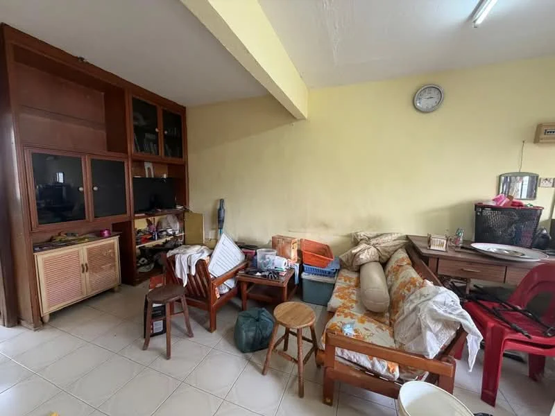 Living features: light-colored walls, tiled flooring, wooden furniture, sofa with floral pattern, wooden chairs, small wooden stools, desk with chair, wall clock, shelves with storage boxes, cluttered space.