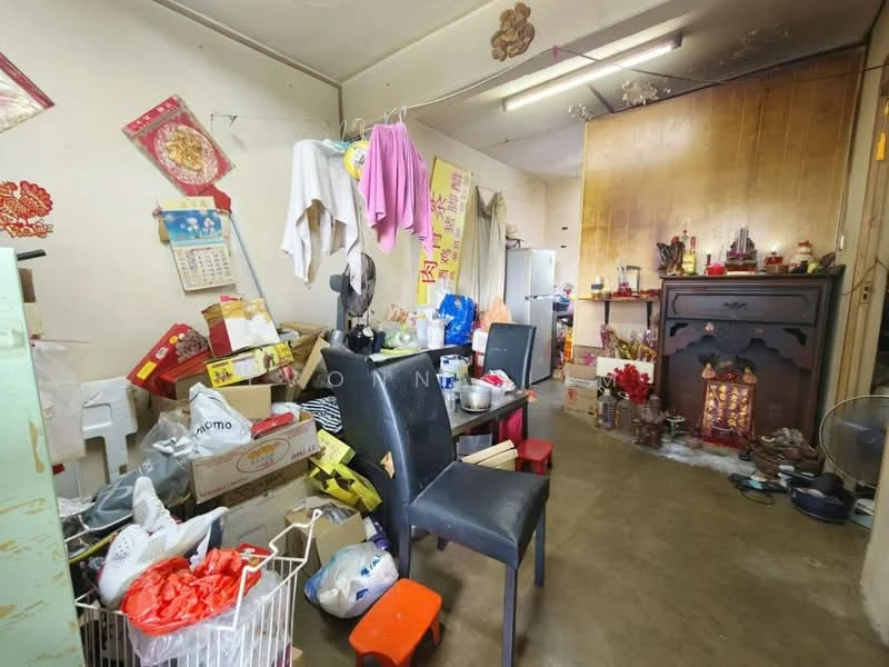 Room features: concrete flooring, wood paneling on walls, various decorations on walls, cluttered space with boxes and items, two black chairs, small wooden table, fireplace with decorative elements, multiple bags and containers, calendar on wall, clothes hanging on wall, various items on shelves.