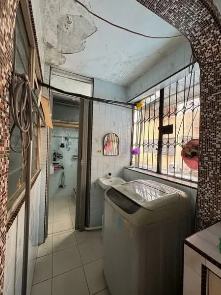 Laundry features: washing machine, white tiled flooring, grey tiled walls, mirror on wall, window with metal grille, sink with faucet, exposed plumbing, light fixture, corner shelving, decorative mosaic border.