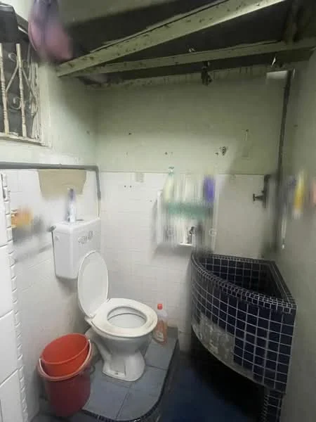 Bathroom features: white toilet, blue tiled flooring, white tiled walls, sink with faucet, metal shelving, red bucket, exposed ceiling beams, window with security bars.