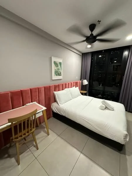 Bedroom features: king-size bed with white bedding, pink upholstered headboard, ceiling fan with 3 blades, wooden study desk, wooden chair, framed artwork on wall, floor-to-ceiling windows, grey curtains, tile flooring, nightstand with lamp; style: modern; view: city.