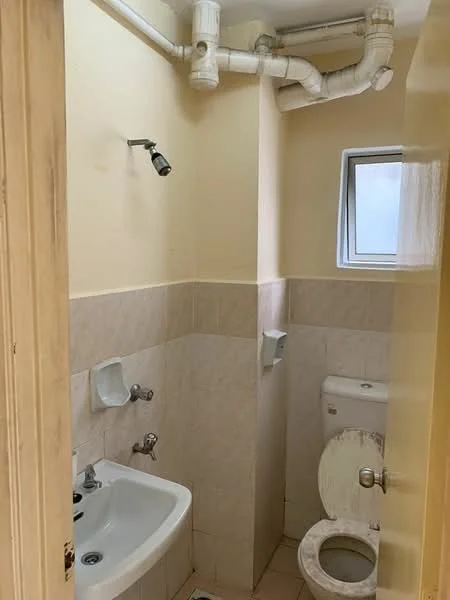 Bathroom features: white toilet, single-sink basin, chrome faucet, wall-mounted mirror, tile flooring, light beige painted walls, window for natural light, exposed plumbing, soap dispenser, toilet paper holder.