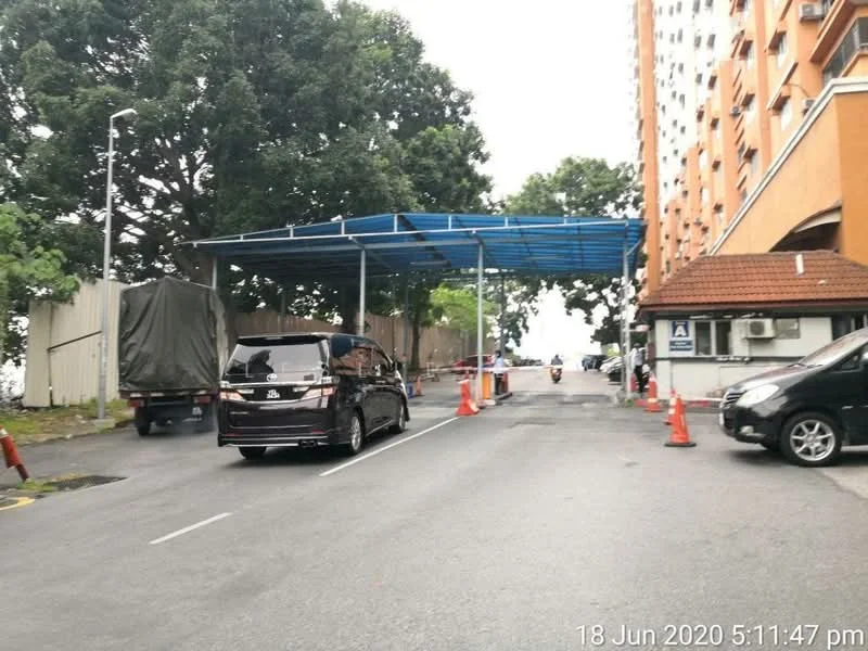 Exterior type: low-rise apartment block; features: covered parking area, paved driveway, traffic cones, surrounding greenery, adjacent to main road, visible vehicle, building facade with orange paint, air conditioning unit on wall; style: modern.