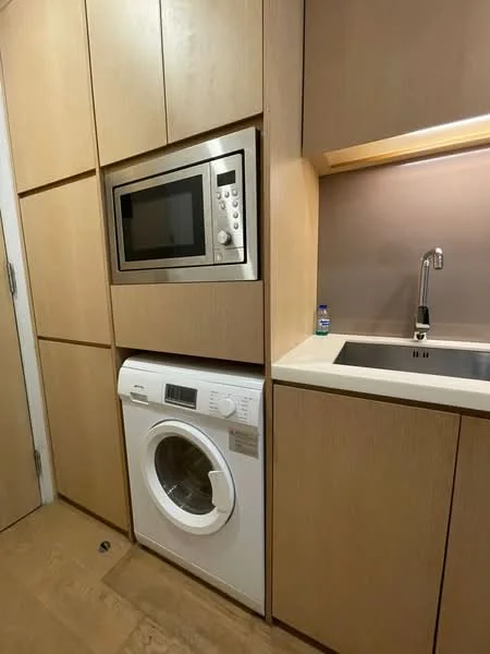Laundry features: built-in cabinetry with wood finish, white washing machine, stainless-steel microwave, single-bowl stainless-steel sink, chrome pull-down faucet, light-colored countertop, cream painted walls, vinyl plank flooring; style: modern.