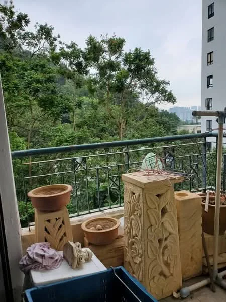 Room features: balcony with railing, potted plants, decorative stone planters, clothes drying rack, blue plastic storage box, textile item on surface, greenery view, urban skyline in distance; view: garden.