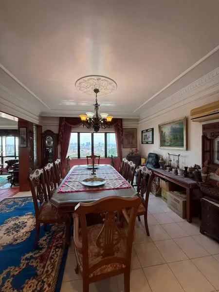 Dining features: rectangular dining table with 12 chairs, chandelier with multiple bulbs, white painted ceiling with decorative rose, cream painted walls with crown moulding, large windows with city view, wooden sideboard, framed artwork on walls, area rug with floral pattern, wooden flooring, air conditioning unit; style: traditional; view: city.