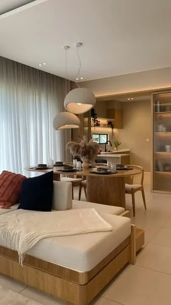 Living features: sofa with light fabric upholstery, wooden coffee table, dining table with wooden finish, 4 dining chairs, pendant lighting with three globes, floor-to-ceiling windows with sheer curtains, decorative vase with dried flowers, neutral wall color, tile flooring, built-in cabinetry with glass doors, kitchen area visible in background; style: modern.