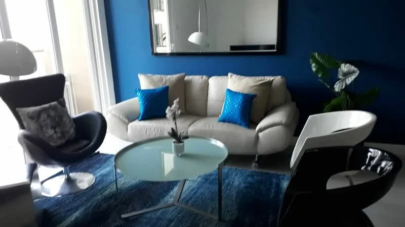 Living features: beige fabric sofa, 2 blue decorative cushions, glass coffee table, blue area rug, black accent chair, white modern chair, large wall mirror, indirect lighting from ceiling, potted plant; style: modern.