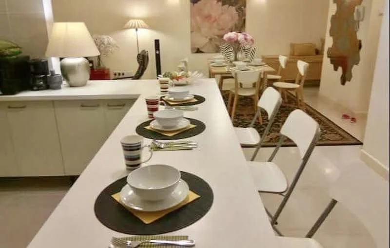 Kitchen features: white cabinetry, marble countertop, stainless-steel sink, chrome faucet, 4 dining chairs, dining table with white surface, decorative wall art, table setting with plates and cutlery, pendant lighting, area rug with geometric pattern, laminate flooring, wall-mounted decor, greenery in decorative pot; style: modern.