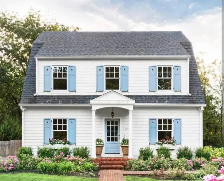 Exterior type: detached bungalow; features: white painted facade, blue shutters, gabled roof with dark shingles, front porch with columns, brick pathway, landscaped garden with flowers, mature greenery, symmetrical window arrangement; style: traditional.