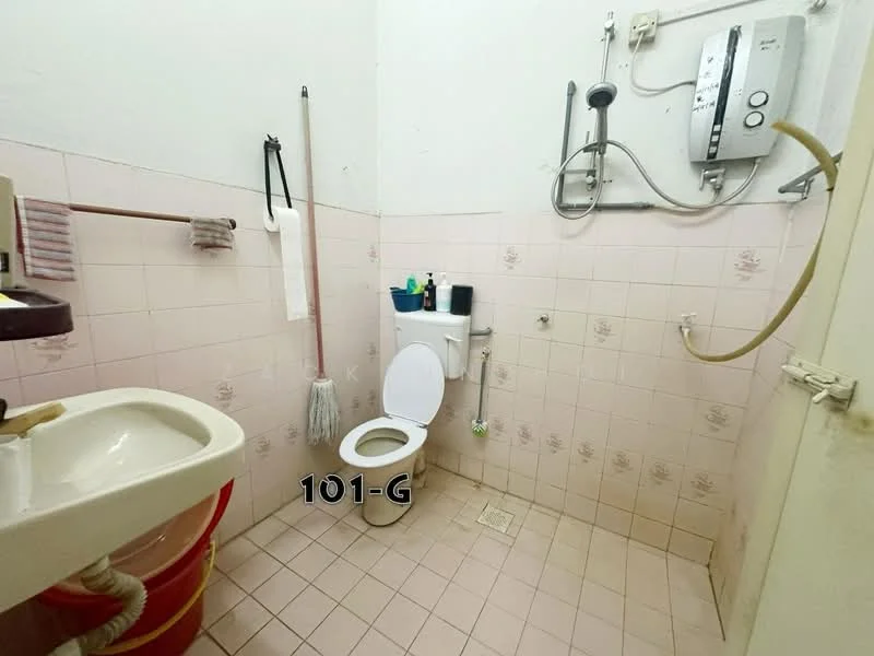 Bathroom features: white ceramic toilet, wall-mounted shower with handheld showerhead, white sink with cabinet, pink tiled walls, pink tiled flooring, water heater mounted on wall, toilet paper holder, cleaning supplies on shelf.